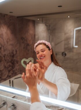 Eine Frau im Bademantel lächelt, während sie sich im Spiegel im Hotel Theresa im Zillertal betrachtet